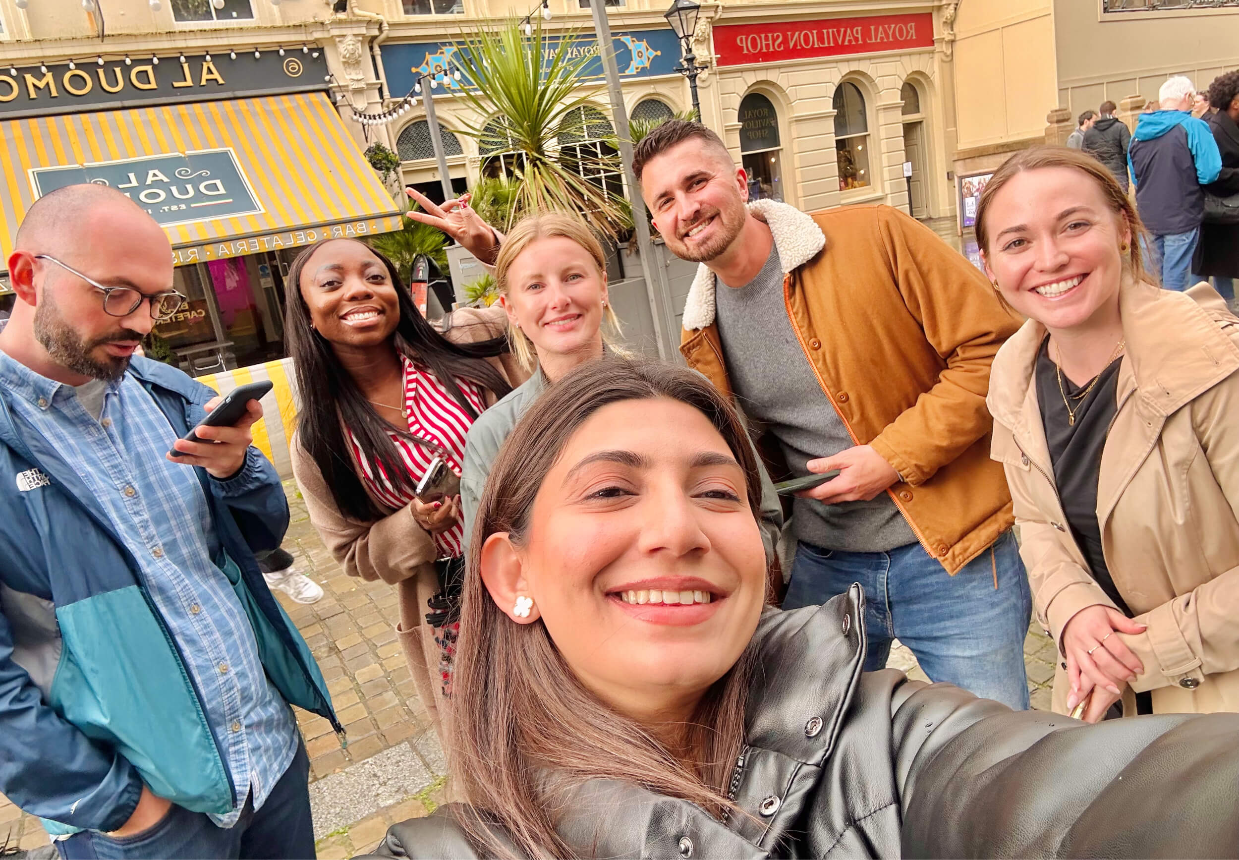 A group of six people taking a selfie playing their treasure hunt in a busy city square, with cafes and shops in the background.